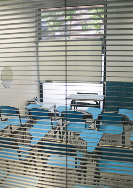 Vista desde fuera de aula con persiana veneciana y pupitres de color azul claro
