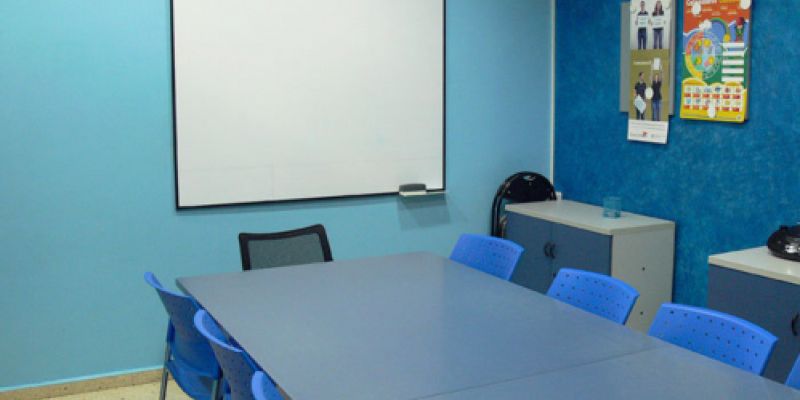 Interior d'aula amb parets de color blau i taula gran al centre i pantalla al fons