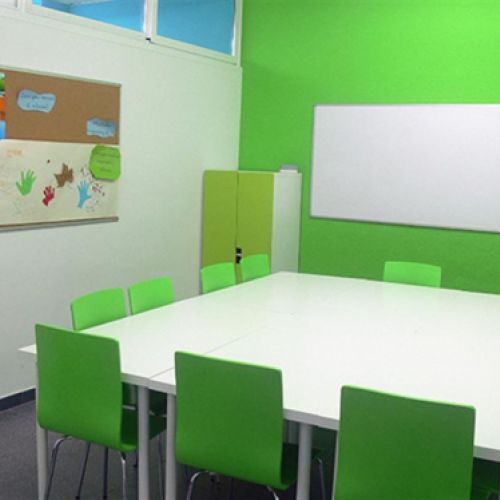Interior d'aula infantil amb taules blanques juntes al centre i cadires i paret de color verd