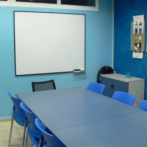 Interior d'aula amb parets de color blau i taula gran al centre i pantalla al fons