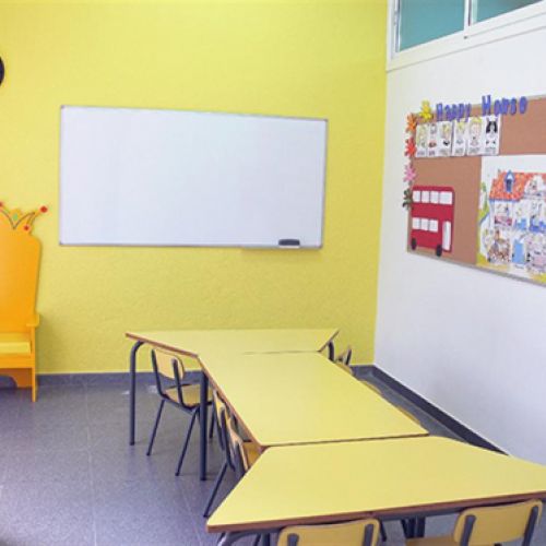 Interior d'aula infantil amb taules i cadires baixes de color groc i tron ​​a paret