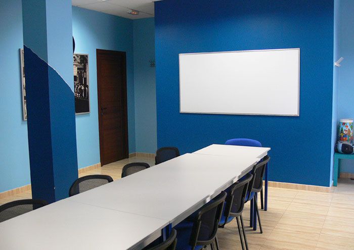 Interior de aula con mesa blanca larga en el centro, paredes de color azul y pizarra blanca en pared