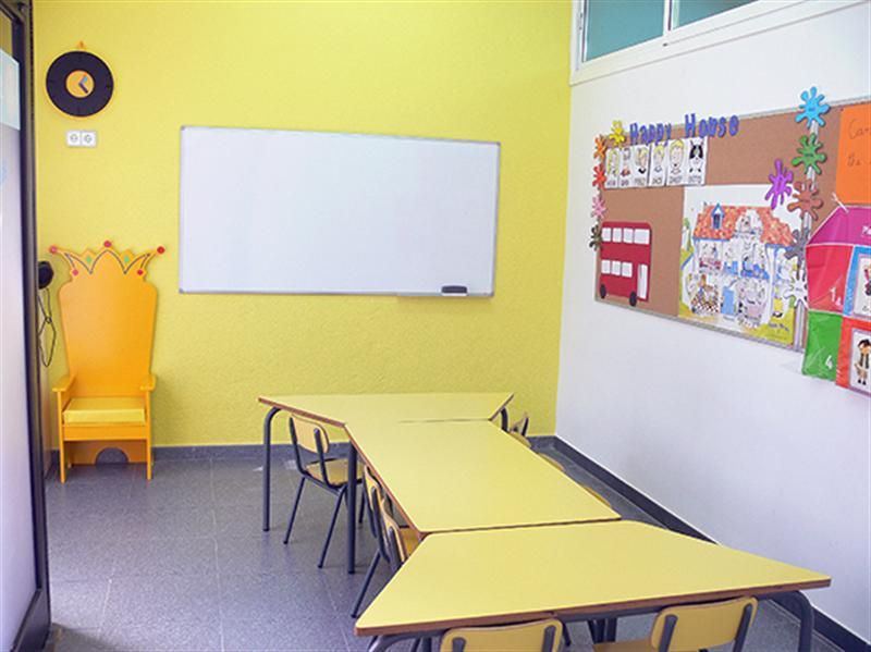 Interior d'aula infantil amb taules i cadires baixes de color groc i tron ​​a paret