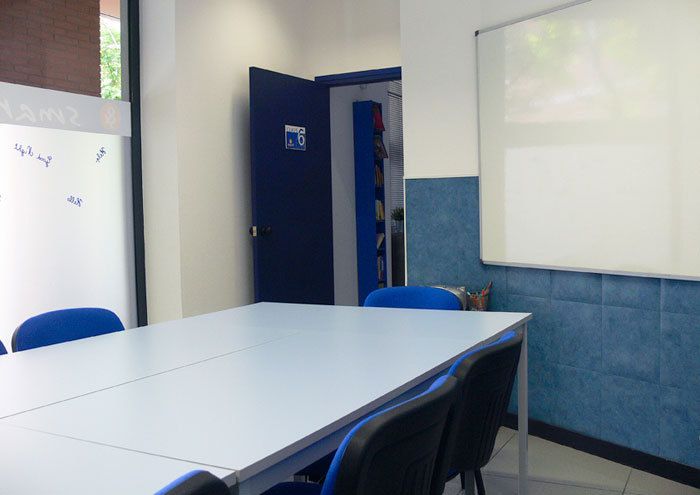 Interior de aula con mesas juntas en el centro, sillas azules a los lados y pantalla blanca en la pared