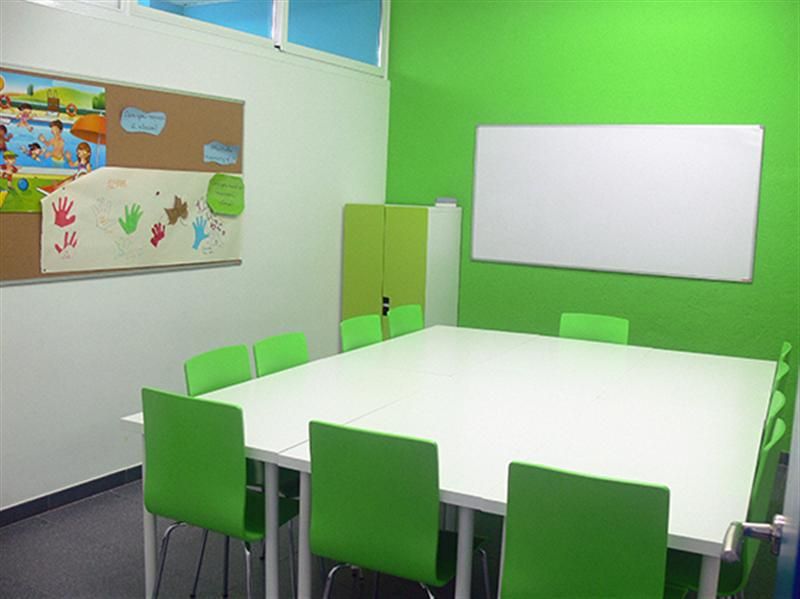 Interior de aula infantil con mesas blancas juntas en el centro y sillas y pared de color verde