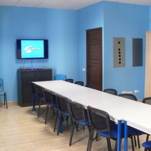 Interior d'aula amb taula llarga al centre, parets de color blau i pantalla digital a paret de fons