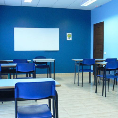 Interior d'aula amb taules, cadires i parets de color blau i pissarra blanca a paret de fons