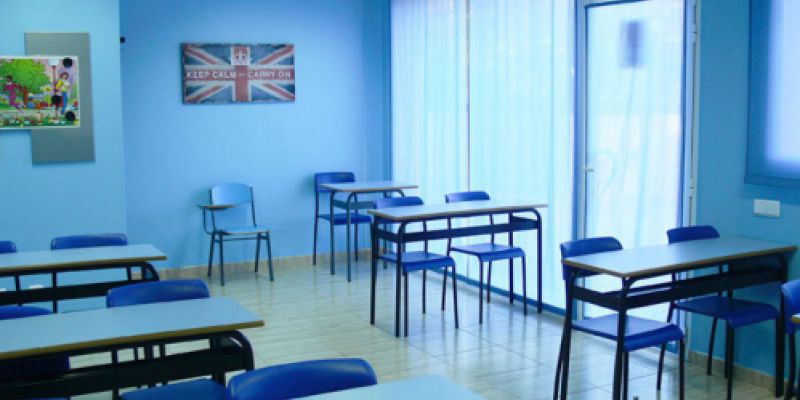 Interior d'aula amb taules, cadires i parets de color blau i quadre amb bandera anglesa a paret de fons
