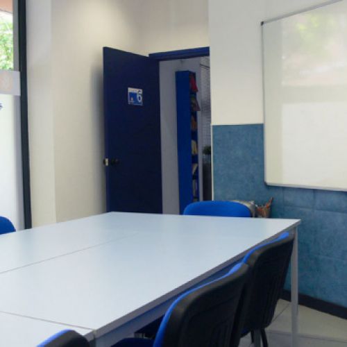 Interior de aula con mesas juntas en el centro, sillas azules a los lados y pantalla blanca en la pared