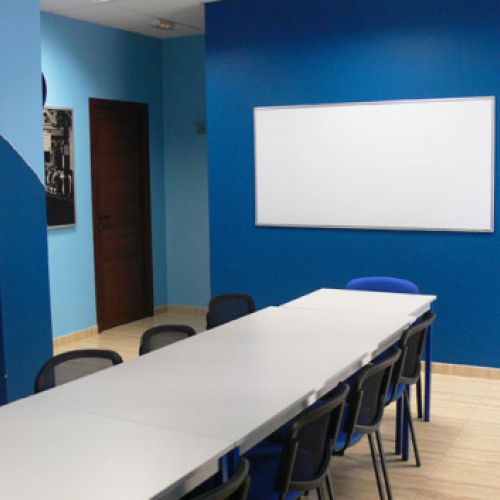 Interior de aula con mesa blanca larga en el centro, paredes de color azul y pizarra blanca en pared