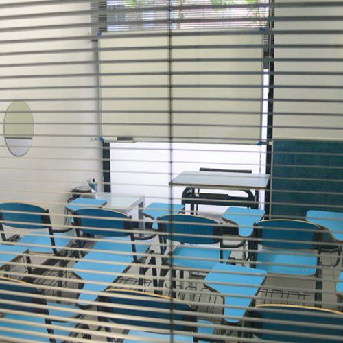 Vista des de fora d'aula amb persiana veneciana i pupitres de color blau clar