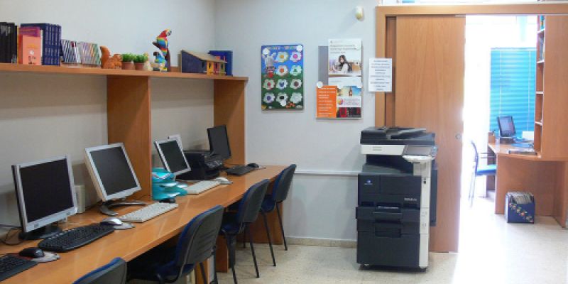 Interior d'aula amb taules juntes i ordinadors a un lateral