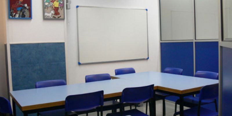 Interior d'aula amb taules a L, cadires blaves als costats i quadres de Tintín a la paret