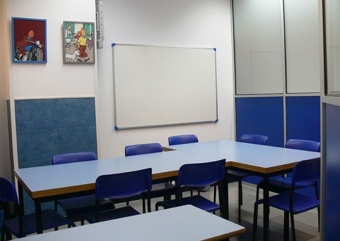 Interior d'aula amb taules a L, cadires blaves als costats i quadres de Tintín a la paret