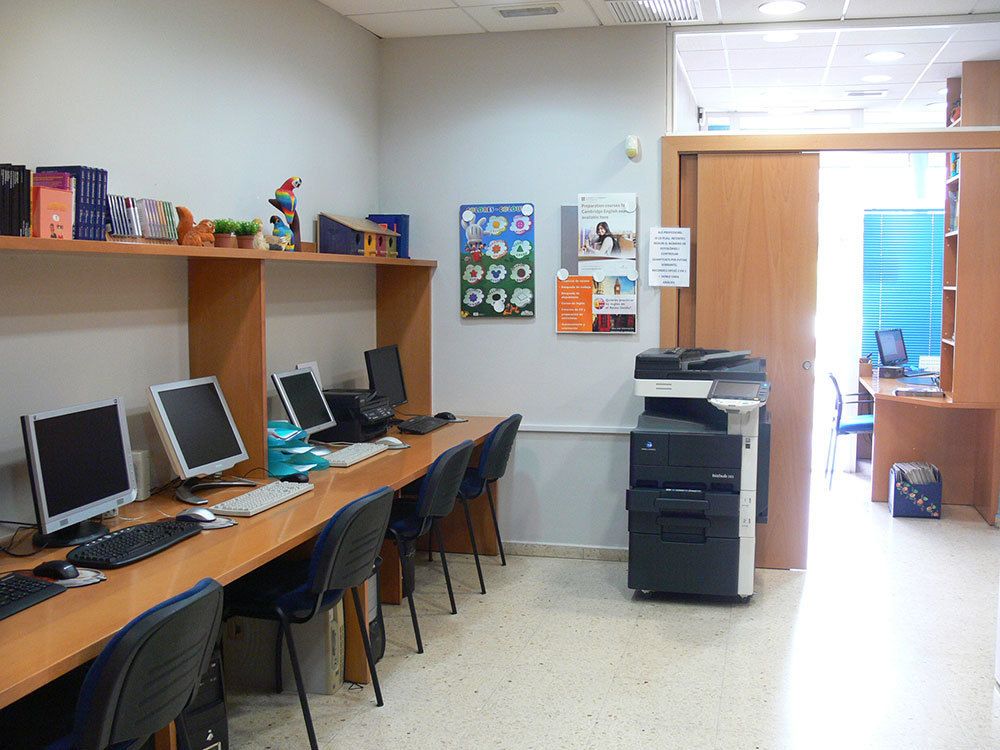 Interior d'aula amb taules juntes i ordinadors a un lateral