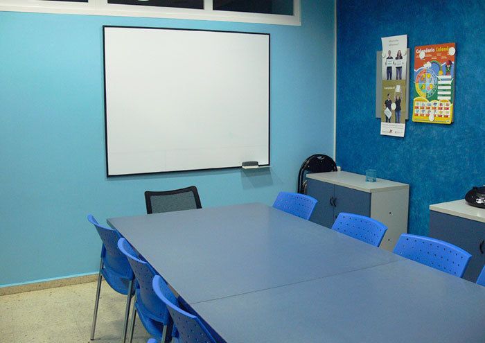 Interior d'aula amb parets de color blau i taula gran al centre i pantalla al fons