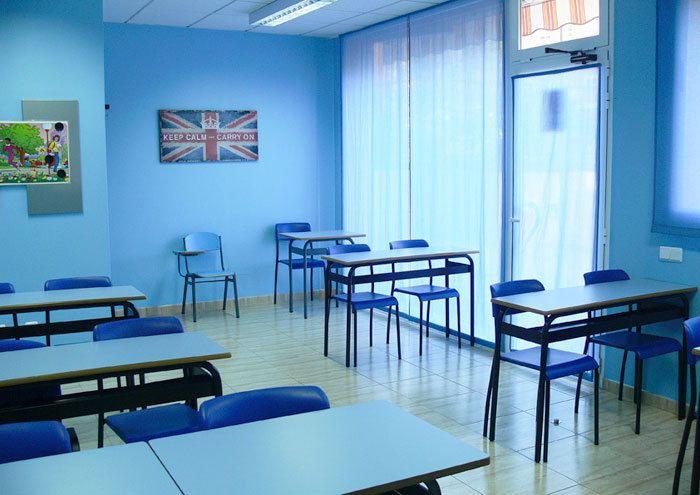 Interior d'aula amb taules, cadires i parets de color blau i quadre amb bandera anglesa a paret de fons