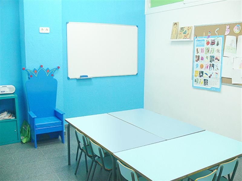 Interior d'aula infantil amb taules i cadires baixes de color blau i tron ​​a paret