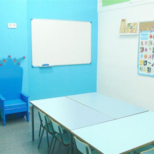 Interior d'aula infantil amb taules i cadires baixes de color blau i tron ​​a paret